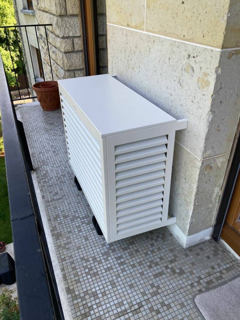 Coffre cache-clim de marque DecoClim, en aluminium blanc, installé sur un balcon. Ce coffre habille joliement un groupe extérieur de climatisation réversible.