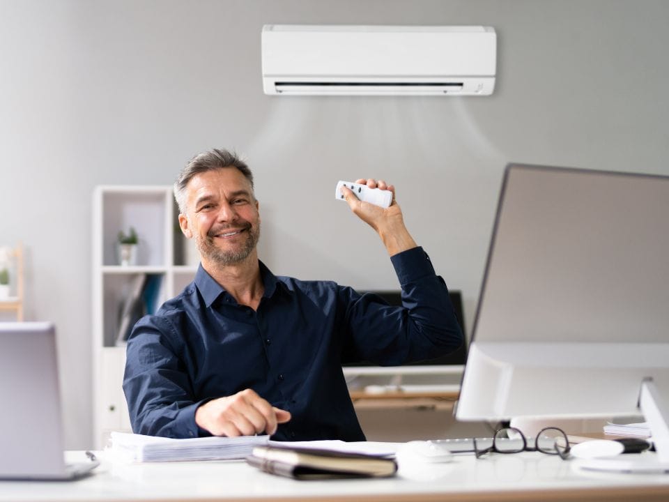 Un homme installé à son bureau et entrain de régler, avec une télécommande, la température de sa climatisation réversible. On voit le mono-split mural à l'arrière plan.