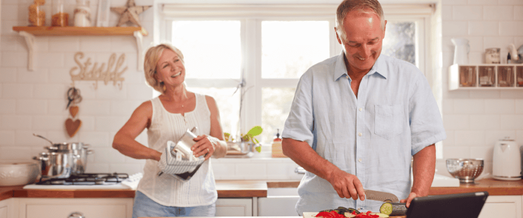 Une femme et un homme debout dans leur cuisine en train de faire à manger. L'homme coupe des légumes et la femme essuie une casserole