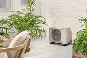 Unité extérieure de climatisation réversible installée sur une terrasse moderne arborée de plantes vertes.