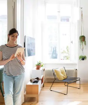 Une femme utilise une tablette pour régler à distance le chauffage de son salon lumineux équipé d'un radiateur classique.