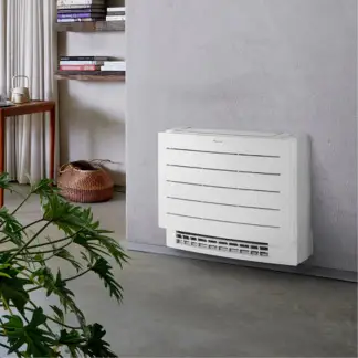 Unité intérieure de climatisation réversible blanche installée au sol contre un mur, dans un intérieur lumineux avec plante et étagères en bois.
